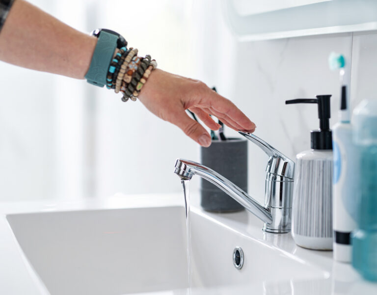 Hand reaching toward metal sink faucet to start water flow in bright modern bathroom
